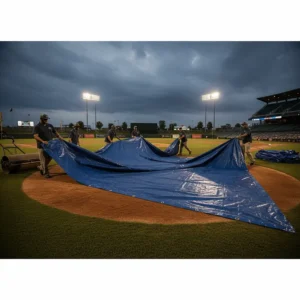 A large, blue baseball field tarp being rolled out by a grounds crew to protect the infield from rain.
